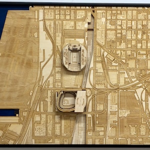 May include: A detailed wooden model of a city skyline, featuring a stadium and a baseball field. The model is made of light brown wood and is set against a dark blue background.