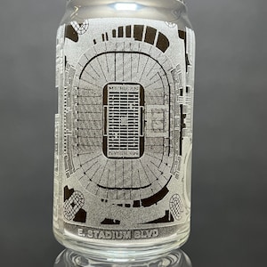 May include: A clear glass can with an etched design of a football stadium. The stadium is labeled "Michigan" and "E. Stadium Blvd."