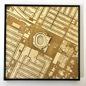 May include: A laser-cut wooden map of a city with a stadium in the center. The map is framed in black and has a light brown wood finish.