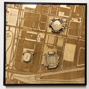 May include: A wooden map of a city, framed in black. The map features detailed laser-cut representations of buildings, streets, and landmarks. The text on the map includes street names and the phrase "DELAWARE EXPY". The overall color is a natural wood tone.