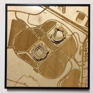May include: A laser-cut wooden map of a sports stadium complex. The map shows the stadium, parking lots, and surrounding roads. The map is framed in black.