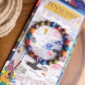 May include: A colorful beaded bracelet with a silver charm that says "Tennessee". The beads are decorated with images of Tennessee landmarks and symbols, including a guitar, a bear, and a state flag. The bracelet is laid on a map of Tennessee.