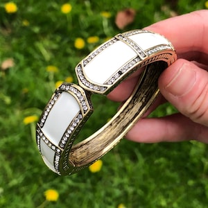 May include: A white enamel and gold tone bangle bracelet with clear rhinestones. The bracelet is open and has a hinged closure.
