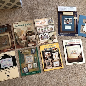 May include: A collection of cross-stitch pattern books and framed artwork, featuring Southwestern themes. The books display various designs, including images of cacti, Native American figures, and landscapes. The color palette includes earth tones, blues, and greens.
