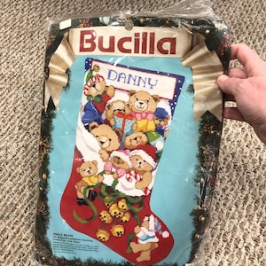 May include: A Bucilla Jingle Bears 16-inch needlepoint stocking kit in a plastic package. The kit features a light blue stocking with a red toe and heel, decorated with multiple teddy bears, gold bells, and a white cuff with the name DANNY.