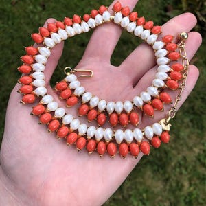 May include: A vintage necklace featuring alternating rows of white faux pearls and orange-red beads. The necklace has a gold-tone metal setting and a hook clasp closure. The design is symmetrical and the beads are evenly spaced.