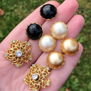 May include: Assortment of decorative buttons in gold, black, and white. The buttons are round and have a raised design. Two of the buttons are ornate with a floral design and clear rhinestones.