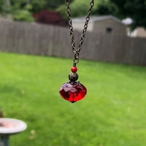 May include: A necklace featuring a bronze-toned metal chain and a pendant composed of a faceted, translucent red glass bead. The pendant is accented with a small, round red bead and a decorative bronze-toned metal bead cap at the top.