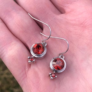 May include: A pair of silver earrings featuring large, round, red gemstones set in a bezel setting. Each earring has a smaller, square-shaped red gemstone dangling below the main stone. The earrings are displayed against a light background.