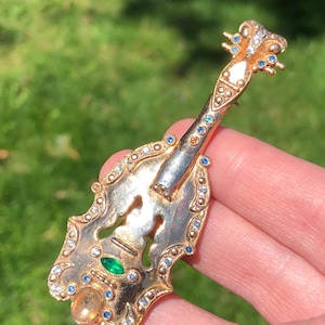 May include: A gold-toned brooch shaped like a violin with blue and clear rhinestones and a green gemstone.