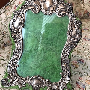 May include: Ornate silver-toned picture frame with a green velvet backing. The frame has a detailed, scrolling design and a curved shape. The frame is designed to hold a photograph or artwork.