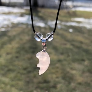 May include: A necklace with a black cord and a light pink, heart-shaped pendant. The pendant is made of a translucent material and is attached to a silver-colored bail. Two clear, round beads are positioned above the pendant.