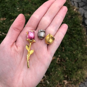 May include: Three gold-toned rose brooches. One rose is pink, one is silver, and one is gold.