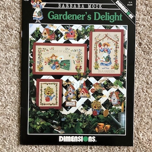 May include: A Barbara Mock Gardener's Delight cross stitch pattern booklet featuring multiple designs of garden scenes, birdhouses, and floral motifs. The cover displays finished projects with text including Sow Seeds of Kindness and Love Grows in a Garden.