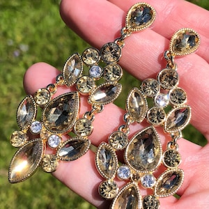 May include: Gold-toned chandelier earrings with a cluster of teardrop, oval, and round-cut crystals. The crystals are in shades of amber, champagne, and clear, creating a sparkling effect. The earrings are designed with a delicate chain.