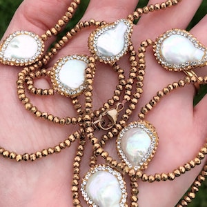 May include: A gold-toned necklace featuring teardrop-shaped, white pearl pendants with crystal accents. The necklace has a beaded chain and a gold clasp. The jewelry is displayed against a soft background.