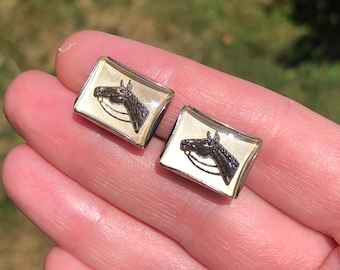 May include: A pair of rectangular cufflinks with a horse head design. The horse head is black with a gold-colored bridle, set against a cream-colored background. The cufflinks have a gold-colored border.