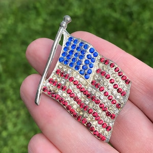 May include: A silver-toned brooch in the shape of an American flag. The flag is made of red, white, and blue rhinestones.