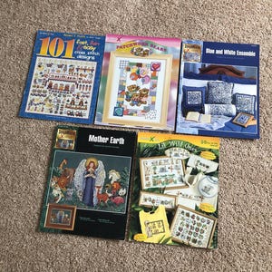 May include: A collection of cross-stitch design books. Titles include "101 Fast, Fun & Easy Cross Stitch Designs", "Patchwork Bears", "Blue and White Ensemble", "Mother Earth", and "Lil Wild Ones". The books feature colorful designs and patterns.