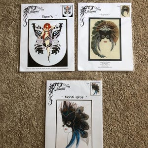 May include: Three cross stitch pattern booklets titled Tigerfly, Papillon, and Mardi Gras. Each booklet features a central illustration of a figure with wings or a mask, enclosed in a clear plastic sleeve with the Illusions brand logo at the top.