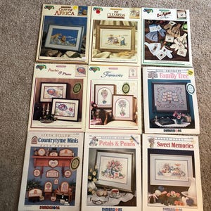 May include: Nine vintage cross-stitch pattern books are displayed, featuring various designs. Titles include "Out of Africa," "The Cleaners," "Bee Appetit," "Peaches & Plums," "Topiaries," "Family Tree," "Countrytyme Minis," "Petals & Pearls," and "Sweet Memories." The books showcase framed needlework.