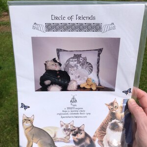 May include: A cross-stitch pattern titled "Circle of Friends" featuring various cat illustrations. The design includes a central image of cats on a pillow, surrounded by other cat images. The pattern is in a clear plastic sleeve.
