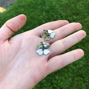 May include: A pair of silver clover-shaped earrings with a small, clear stone in the center of each clover.