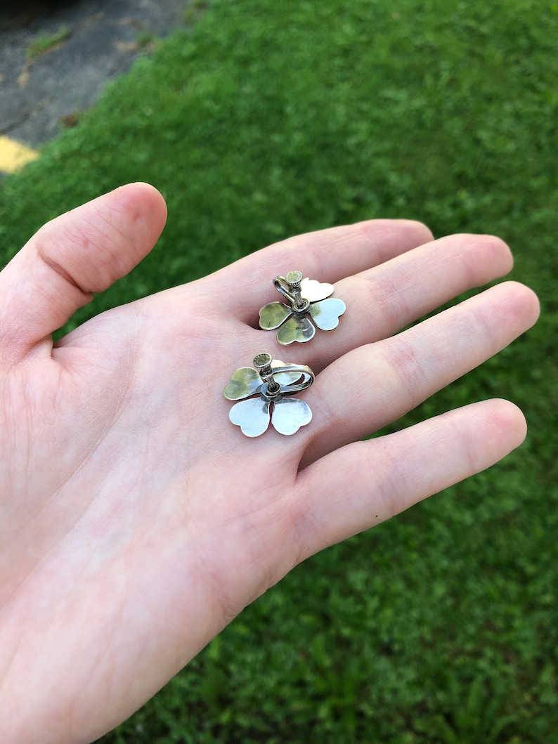 May include: A pair of silver clover-shaped earrings with a small, clear stone in the center of each clover.