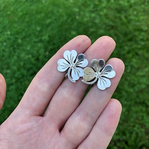 May include: Two silver four-leaf clover charms, one slightly larger than the other, with intricate details, held in a hand against a green grass background.