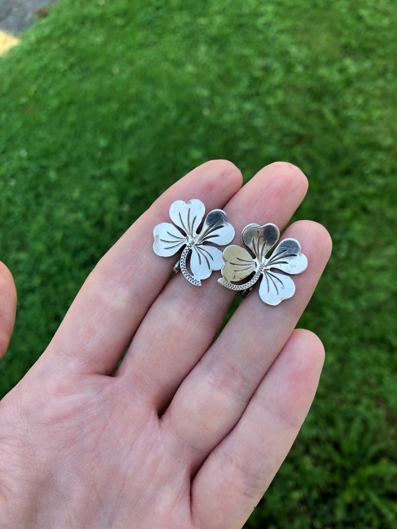 May include: Two silver four-leaf clover charms, one slightly larger than the other, with intricate details, held in a hand against a green grass background.