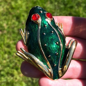 May include: A green frog-shaped brooch with gold-colored accents. The frog has red eyes and small gold dots on its back. The brooch is made of a shiny, metallic material.