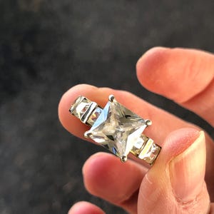 May include: A silver ring with a large, square, clear gemstone as the focal point. The ring has smaller rectangular stones flanking the main stone. The ring is held between fingers.