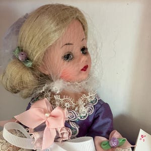 May include: A porcelain doll with blonde hair, wearing a purple dress with lace trim and a pink bow. The doll has a veil and a flower in her hair.