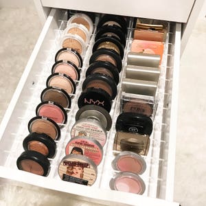 May include: A white drawer organiser with clear dividers holds a variety of round makeup compacts. The compacts are in various shades of brown, pink, and black. Some of the compacts have text on them, including "NYX", "CINDY-LOU MANIZER", and "BETTY LOU MANIZER".