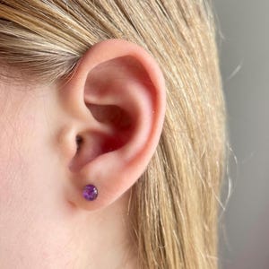 May include: Close-up of a person's ear with a small, round, purple stud earring. The earring is set in a simple, minimalist design. The person has blonde hair.
