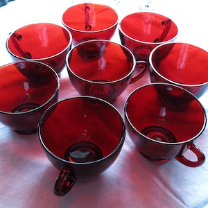 May include: A set of eight matching ruby red glass teacups arranged in a circular pattern. Each cup features a smooth, translucent finish, a rounded bowl shape, and a small, curved handle attached to the side for holding the beverage container.