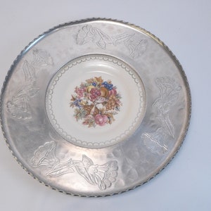 May include: A silver serving tray with an embossed floral design around the edge. The center of the tray features a white porcelain plate with a colorful floral and fruit design.
