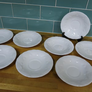 以下が含まれることがあります： 隆起したデザインが施された白いセラミック製のソーサー8枚セット。ソーサーは木製の表面に半円形に配置されています。
