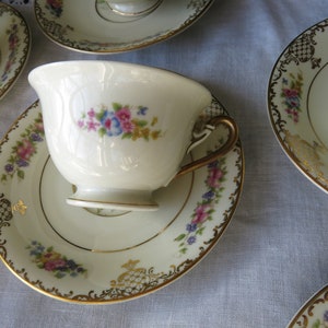 Könnte beinhalten: Eine weiße Teetasse und Untertasse mit floralem Muster und goldener Verzierung. Die Tasse hat ein zartes Blumenmuster in Rosa-, Blau- und Gelbtönen. Die Untertasse hat ein passendes Blumenmuster und einen goldenen Rand.