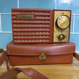 May include: A brown leather carrying case with a vintage brown and gold Silvertone radio inside. The radio has a dial with numbers 5 through 10 and a knob for volume control.