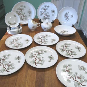May include: A collection of white ceramic dinnerware with a pinecone and branch design. The set includes plates of various sizes, a creamer, a sugar bowl, and saucers. The plates are arranged on a wooden table, showcasing a vintage aesthetic.