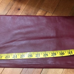 May include: A close-up of a burgundy faux leather fabric with a yellow measuring tape along the bottom edge. The tape reads 21 to 32 inches.