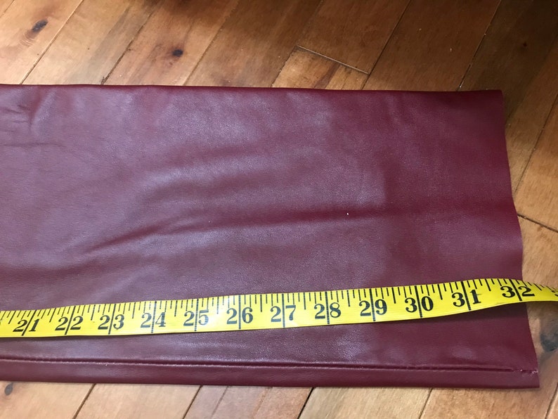 May include: A close-up of a burgundy faux leather fabric with a yellow measuring tape along the bottom edge. The tape reads 21 to 32 inches.
