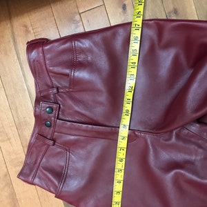 May include: Burgundy leather pants with a zipper fly and button closure. The pants have a seam down the front and a seam down the back. The pants are measured with a yellow tape measure.