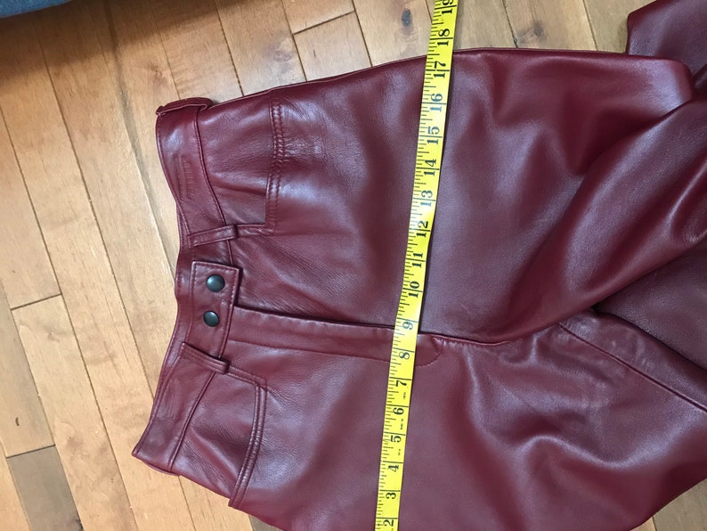 May include: Burgundy leather pants with a zipper fly and button closure. The pants have a seam down the front and a seam down the back. The pants are measured with a yellow tape measure.