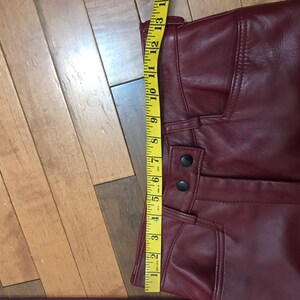 May include: A pair of burgundy leather pants with a zipper fly and button closure. The pants have a seam down the front and a pocket on the right side. A measuring tape is placed along the waistband of the pants.
