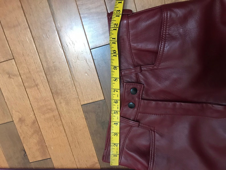 May include: A pair of burgundy leather pants with a zipper fly and button closure. The pants have a seam down the front and a pocket on the right side. A measuring tape is placed along the waistband of the pants.