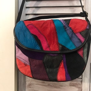 以下が含まれることがあります： 黒いストラップが付いたパッチワークレザーのショルダーバッグ。バッグは赤、青、紫、黒など、さまざまな色の革のピースで作られています。バッグには、黒い革のトリムが付いた湾曲したフラップがあります。