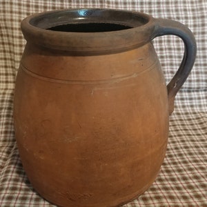May include: A large, brown, earthenware crock with a wide mouth and a single handle. The crock has a dark brown glaze on the rim and handle.