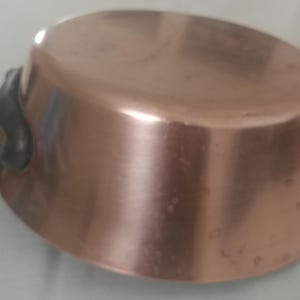 May include: A copper saucepan with a dark handle. The pot has a rounded base and a shiny, reflective surface. The handle is attached to the side of the pot. The saucepan is likely used for cooking.